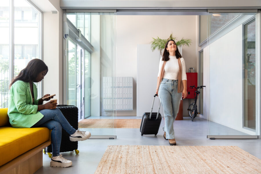 Happy Young Caucasian Woman Enters Hotel Reception With Trolley Suitcase. Copy Space